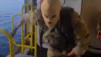 Figura con máscara clara, ojos y boca oscuros, vestida con chaleco y ropa de camuflaje, sube una estructura amarilla con el mar azul visible al fondo