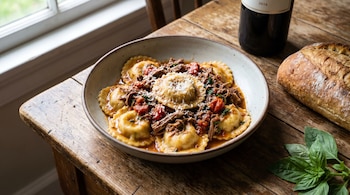 Un plato de ravioles de cordero caseros con salsa roja, carne deshebrada y queso rallado, sobre una mesa de madera. Cerca hay un pan y una botella de vino tinto.