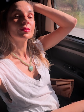 Mujer en coche, top blanco, collar de cuentas verdes con dije de corazón. Luz del sol en rostro. Brazo levantado. Bolso de madera de listones