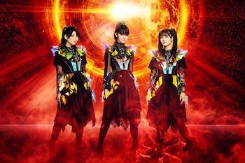 Tres mujeres de Babymetal con elaborados trajes oscuros y detalles iridiscentes posan ante un fondo vibrante de color rojo y naranja intenso