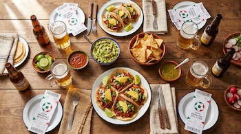 Vista cenital de una mesa de madera con tacos al pastor, guacamole, totopos, salsas y cervezas, acompañada de boletos de la Copa Mundial de la FIFA.