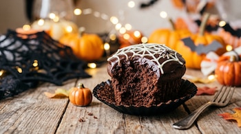 Un pastelillo de chocolate mordido con dibujo de telaraña en una mesa de madera junto a calabazas pequeñas, un tenedor y luces decorativas.