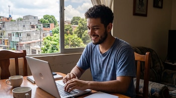 Primer plano de un joven salvadoreño sonriendo y tecleando en su laptop sobre una mesa de madera, con una ventana que muestra un paisaje urbano.