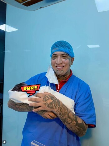 Hombre tatuado con gorro quirúrgico azul y mascarilla colgando, sonríe mientras sostiene a un bebé envuelto. Fondo azul claro