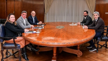 Cuatro hombres y dos mujeres están sentados alrededor de una gran mesa de madera pulida en una sala formal con cortinas claras y paneles de madera