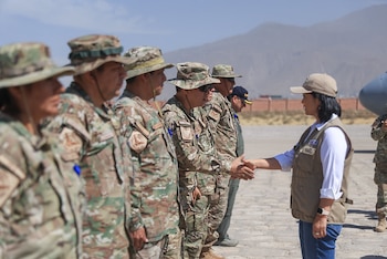 Una mujer con gorra y chaleco saluda de mano a una fila de personas con uniforme de camuflaje en una explanada al aire libre