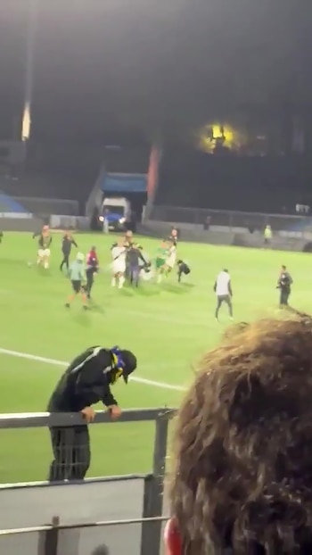 Varios espectadores y futbolistas corren sobre el césped de un estadio iluminado de noche tras una valla metálica