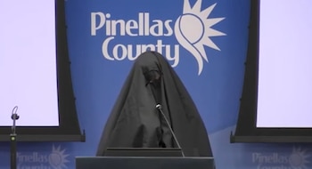 Una persona cubierta por una tela negra frente a un podio con micrófono, ante un fondo azul con el texto y logotipo de Pinellas County