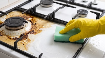 Una mano con guante amarillo sostiene un estropajo verde y limpia una estufa de esmalte blanco con parrillas negras de hierro fundido, que presenta suciedad y espuma.