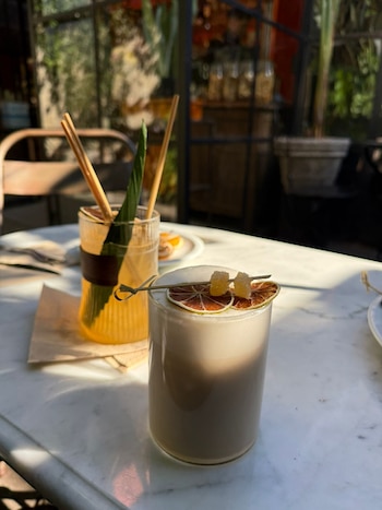 Primer plano de dos bebidas de té sobre una mesa de mármol: una cremosa con rodajas de cítricos y otra clara con garnish de palitos y hoja