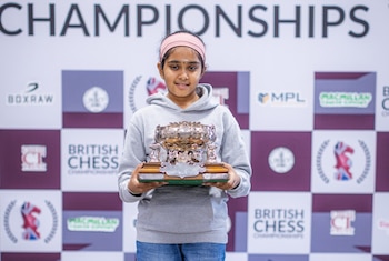 Una niña con sudadera gris y cinta rosa sostiene un trofeo metálico frente a un panel publicitario del campeonato de ajedrez