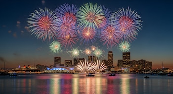 Fuegos artificiales multicolores explotan sobre el horizonte de Long Beach y su puerto al anochecer, reflejándose en el agua, con barcos visibles.
