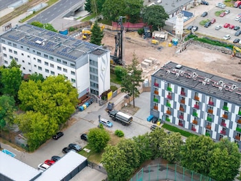 Dos edificios, uno con paneles solares, un sitio de construcción con maquinaria pesada, vehículos, árboles, una carretera y contenedores de basura