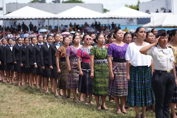 Fila de mujeres en un campo. Al frente, una mujer saluda con uniforme. Detrás, otras visten trajes negros con corbatín azul o atuendos tradicionales coloridos