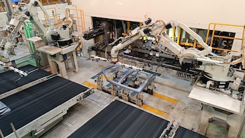 Vista aérea de una línea de ensamblaje automatizada con brazos robóticos gris claro sobre cintas transportadoras negras y una pieza de automóvil metálica