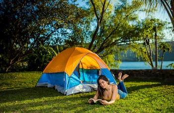 Mujer recostada en el césped leyendo un libro junto a una tienda de campaña naranja y azul, con árboles y una laguna al fondo bajo cielo azul