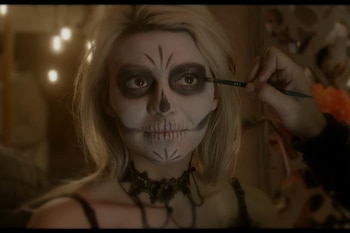 Primer plano de una persona con cabello rubio recibiendo maquillaje de calavera blanco y negro en el rostro, aplicado por una mano con un pincel