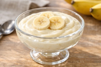 Un postre de crema de plátano con yogur natural y tres rodajas de plátano en un plato de cristal, sobre una mesa de madera bajo luz natural.