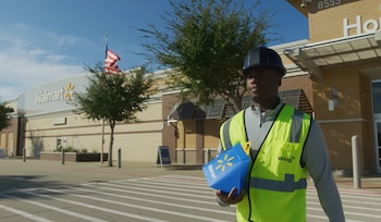 Un hombre con chaleco amarillo y casco oscuro lleva un paquete azul de Walmart. Camina en una acera con rayas frente a la tienda Walmart y árboles