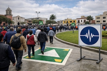 Multitud de personas de espaldas y perfil evacúan una plaza central de ciudad peruana, con edificios, palmeras y señalizaciones verdes en el suelo y un cartel azul del IGP.