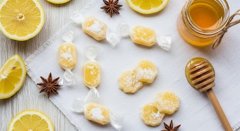 Caramelos de miel y limón, algunos envueltos y otros cubiertos de azúcar, con limones, anís estrellado, un tarro de miel y un dipper, sobre tela blanca.
