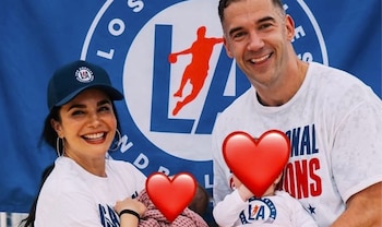 Retrato de Martha Higareda y Lewis Howes sonriendo, cada uno sosteniendo un bebé cubierto por un corazón rojo, frente a un estandarte azul con un logo de Los Ángeles
