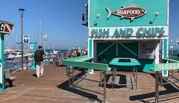 Vista de un muelle de madera con un edificio turquesa que dice 'Fish and Chips' y 'Seafood', mesas y bancos verdes, personas, botes en una marina y cielo azul