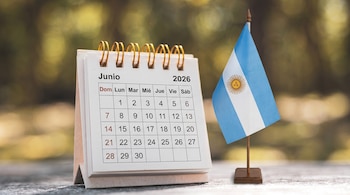 Calendario de escritorio para junio de 2026 junto a una pequeña bandera de Argentina en un mástil de madera, sobre una mesa con fondo verde borroso.