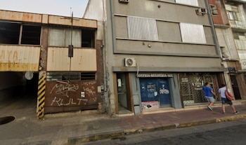 Vista frontal de una calle urbana con una joyería gris a la derecha y una cochera de dos pisos, con puerta de metal oxidado y grafitis, a la izquierda