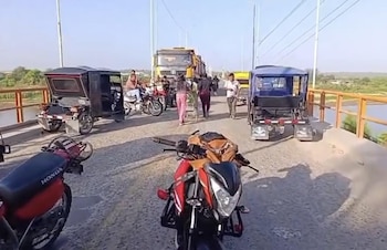 Vista de un puente con personas y varios vehículos, incluyendo motocicletas y mototaxis, bloqueando el paso, bajo un cielo claro y un río al lado