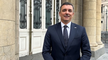 Un hombre de mediana edad sonríe con traje oscuro, camisa blanca, corbata de lunares y pin de YPF, frente a una puerta blanca con rejas y columnas de piedra