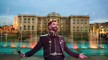 Alejandro Fernández viste traje de charro y sostiene un micrófono frente a fuentes iluminadas de colores y un edificio histórico.