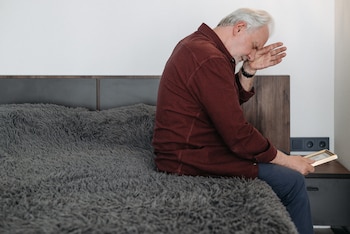 Hombre mayor con camisa roja a cuadros y pantalón azul sentado de perfil en una cama con manta gris. Sostiene un marco de foto dorado y lleva una mano a la cara