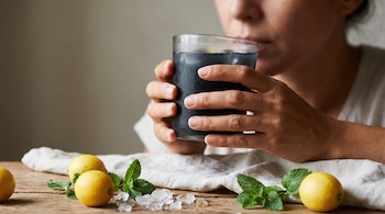 Manos de una persona adulta sostienen un vaso con limonada negra cerca de su boca. Sobre una mesa hay limones, hojas de menta y hielo.