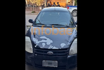 Vista frontal de un automóvil Ford negro con la pintura del capó deteriorada, con agentes de policía y un vehículo policial en el fondo