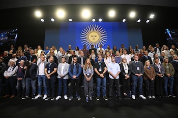 Gran grupo de hombres y mujeres de pie en varias filas, mirando al frente. Detrás, una gran pantalla azul con la bandera argentina y el Sol de Mayo