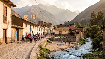Calle empedrada de un pueblo con casas de adobe, personas con trajes típicos, banderas, río con orilla de arena, montañas y cielo con nubes.