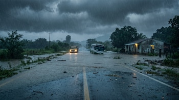 Un camino rural inundado bajo una intensa lluvia, con un autobús blanco y un camión amarillo, ambos en la carretera, y una casa con luces encendidas a la derecha. Cielo nublado.