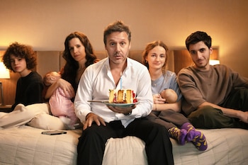 Un grupo de cinco personas y dos bebés sentados en una cama. Un hombre en el centro sostiene un trozo de pastel con una vela encendida