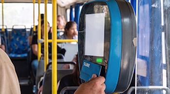 Mano de una persona acercando una tarjeta SUBE a un lector electrónico de color azul y gris dentro de un autobús con pasajeros y asientos azules.