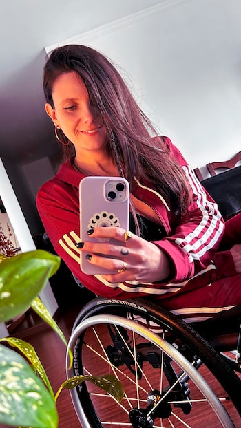 Laura Scianca sonriendo, sentada en una silla de ruedas, vistiendo un chándal rojo con rayas blancas mientras se toma una selfie con su teléfono morado