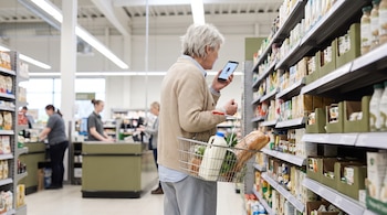 Una persona adulta mayor de cabello plateado corto sostiene un smartphone con un ícono de micrófono en pantalla y una canasta de compras con leche y pan en un supermercado.