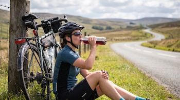 Una mujer con casco y gafas de sol toma de una botella transparente sentada junto a una bicicleta apoyada en un poste.
