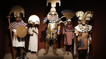 Cinco maniquíes con vestimentas, tocados de metal dorado y cetros ceremoniales mochicas se exhiben sobre pedestales en un museo