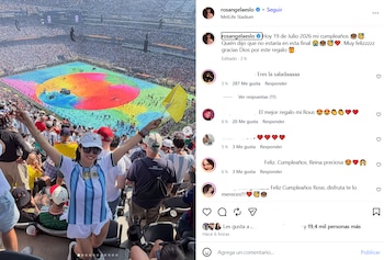 Mujer con camiseta de Argentina, shorts y gorra blanca, gafas de sol y brazos extendidos en un estadio. Campo de juego colorido y bandera amarilla