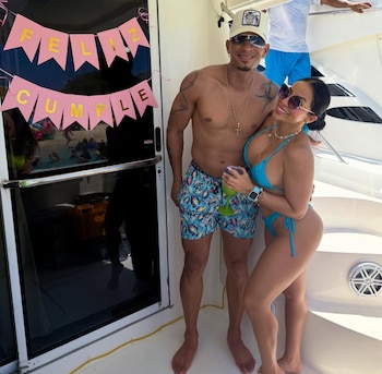 Un hombre y una mujer sonrientes posan en la cubierta de un yate, con un cartel de Feliz Cumple. Hay personas en el agua de fondo