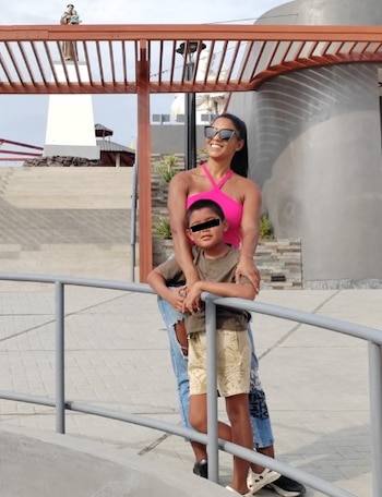 Una mujer con top rosa, jeans y gafas de sol, sonríe abrazando a un niño con camiseta marrón y shorts, apoyados en una barandilla gris al aire libre