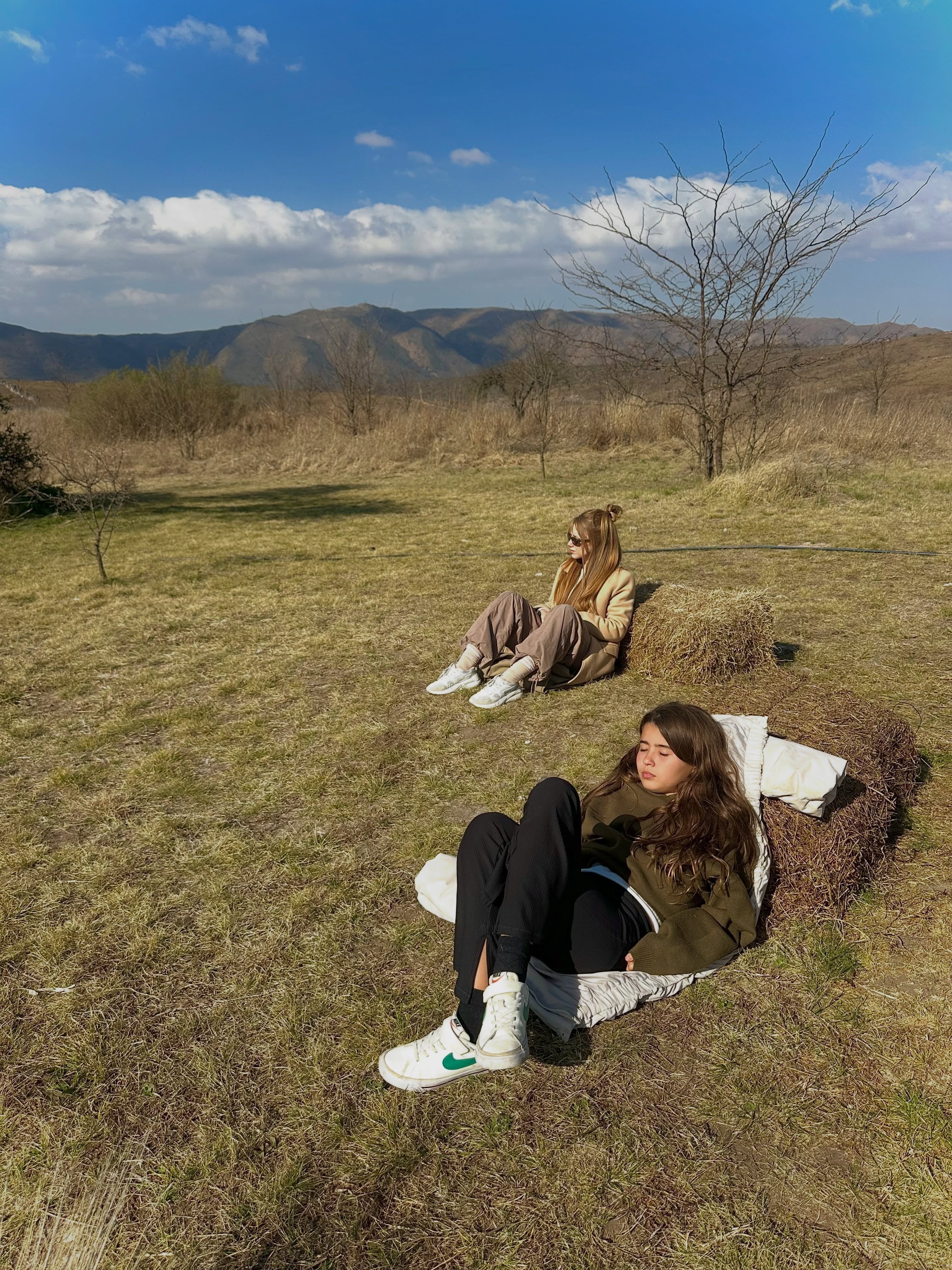 La hija de Nicolás Cabré, Rufina, y Rocío Pardo descansan sobre fardos de heno en un paisaje rural de colinas bajo un cielo nublado.