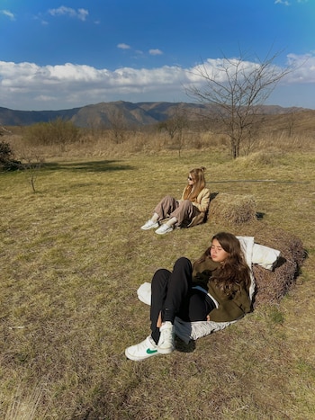 Dos mujeres jóvenes, una acostada y otra sentada, sobre fardos de heno en un campo con hierba seca, árboles desnudos y montañas bajo un cielo azul con nubes