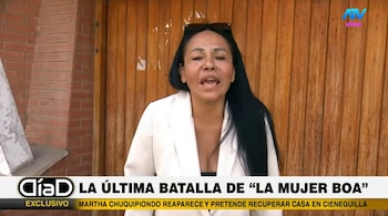 Mujer con cabello largo oscuro, gafas de sol en la cabeza y saco blanco, habla frente a pared de ladrillos y puerta de madera. Logotipos y texto periodístico visibles